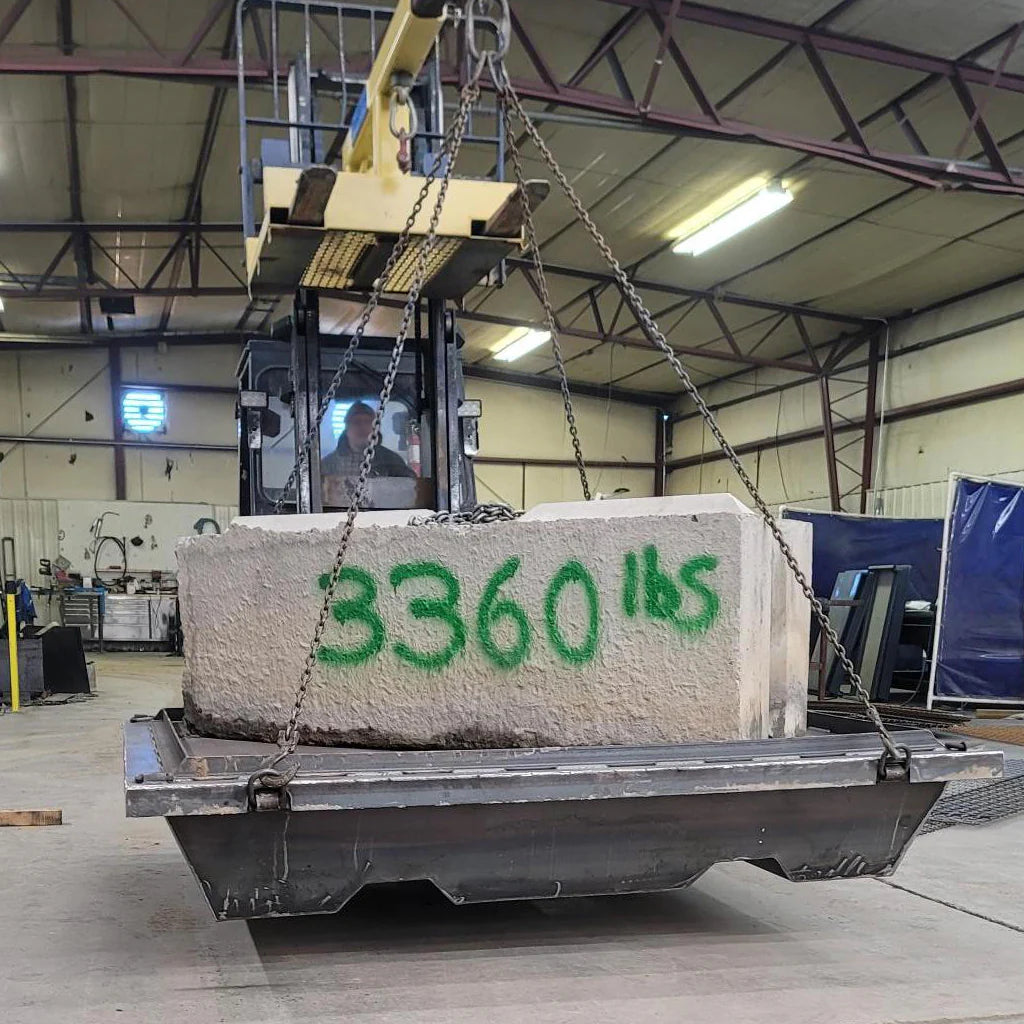 Large concrete block with '3360 lbs' in green spray paint, suspended by a crane in an industrial setting.